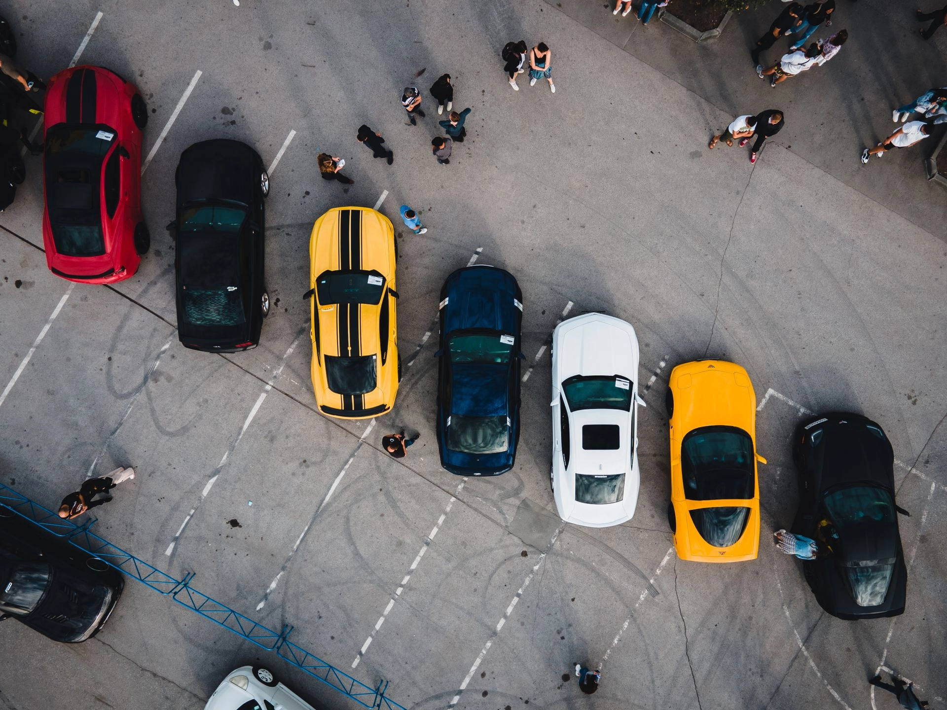 Overhead view of a row of red, black, yellow, blue, white, and orange cars parked on an auction lot with people walking between them.